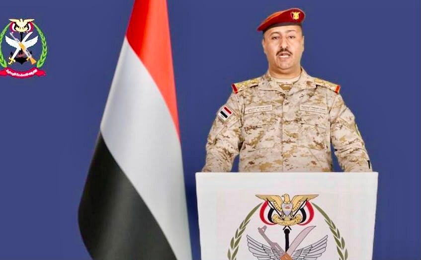 Yemeni armed forces spokesman in uniform speaking at a podium beside the national flag and the armed forces emblem
