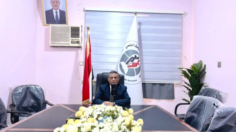 Yemen's minister of higher education and scientific research seated at a desk in his office beneath the national flag during the virtual signing ceremony