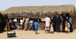 Displaced Yemeni families queue outside a reed shelter in a desert settlement, with water jerrycans and a generator nearby