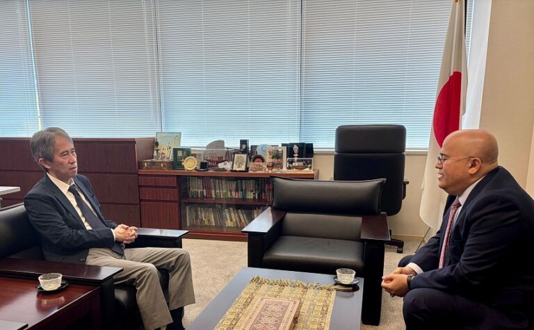 Two diplomats seated facing each other in a Tokyo office with a Japanese flag behind them