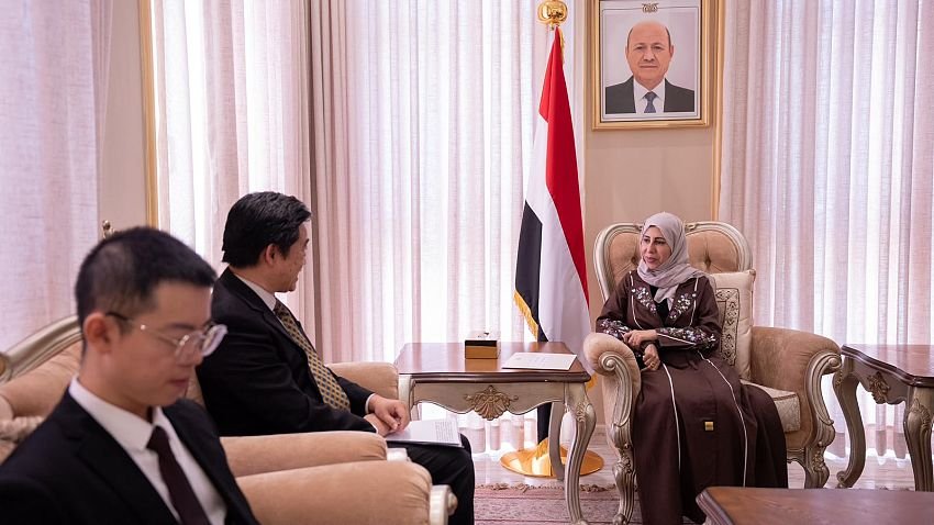 Yemeni Foreign Minister Afrah al-Zouba seated in a meeting room with visiting officials, beside a Yemeni flag and a portrait of Rashad al-Alimi