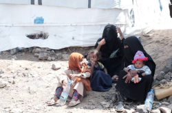 A displaced Yemeni woman and three young children sit on the ground beside a tent shelter