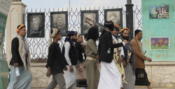 Men in traditional Yemeni dress walk past a wall hung with framed portraits during a funeral procession
