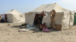 A displaced family shelters beside makeshift tents at a camp in Yemen, with bedding and belongings on the bare ground.