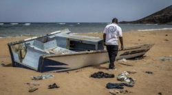 A man walks beside a wrecked wooden boat on a shore scattered with clothing and debris after a migrant vessel sank